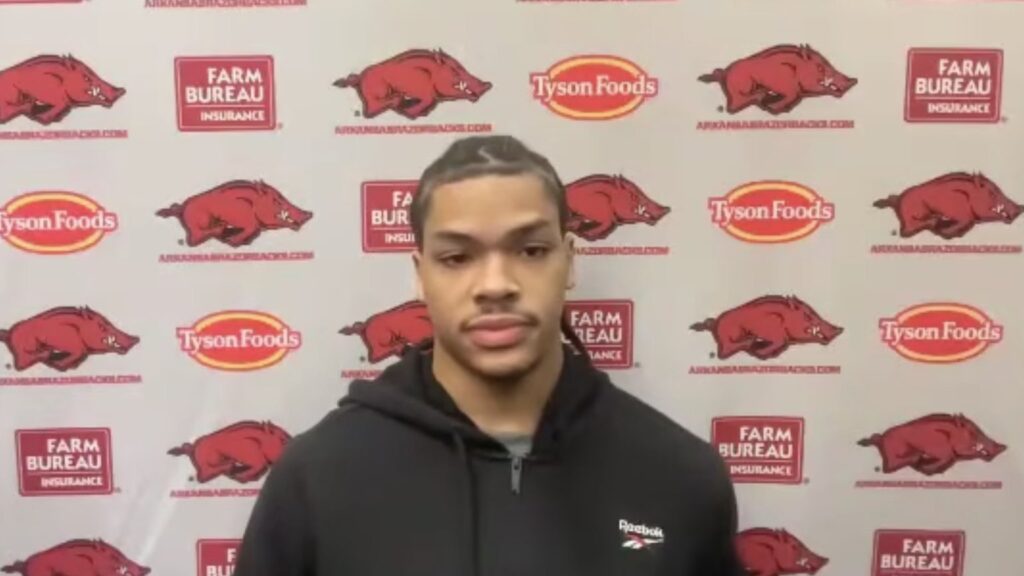 Darius Acuff Jr. in a black hoodie at an Arkansas basketball media session in front of a team logo backdrop