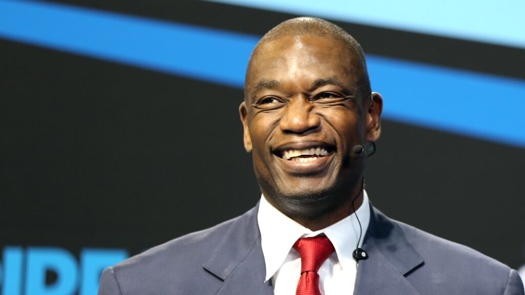 Dikembe Mutombo smiling while speaking on stage in a suit and red tie