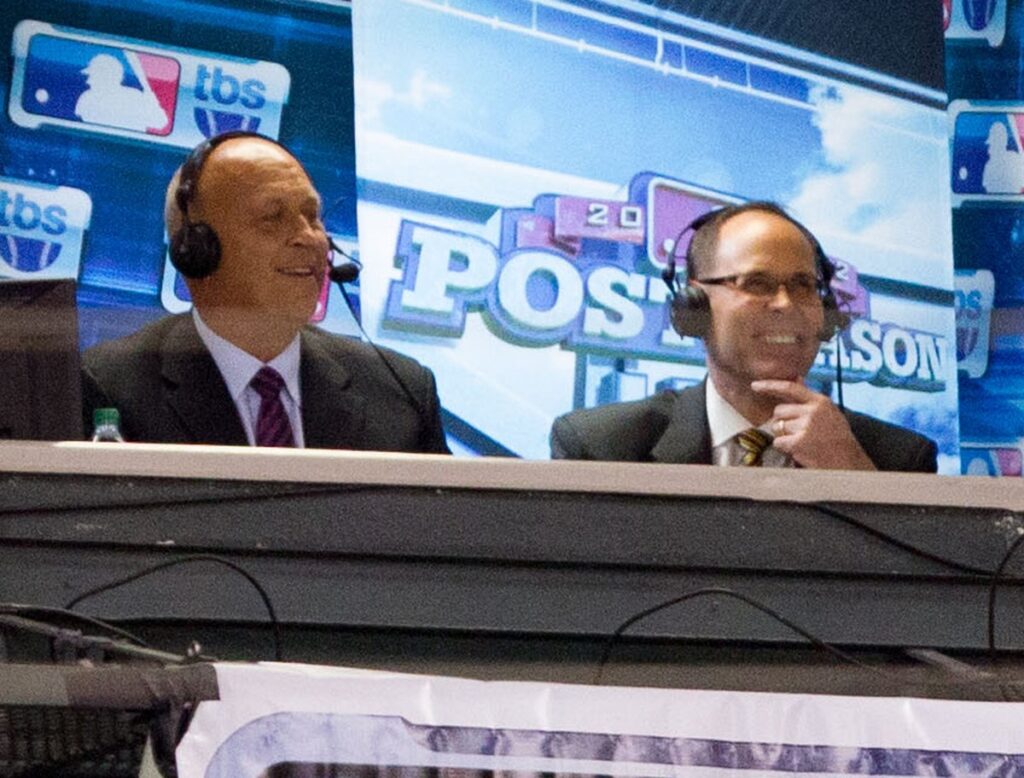 Ernie Johnson smiling in a broadcast booth with headphones on, next to Cal Ripken Jr.