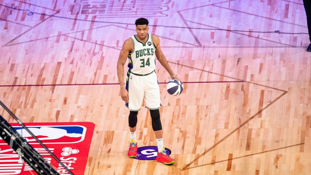 Giannis Antetokounmpo in a white Milwaukee Bucks No. 34 jersey dribbling a basketball on an All-Star court