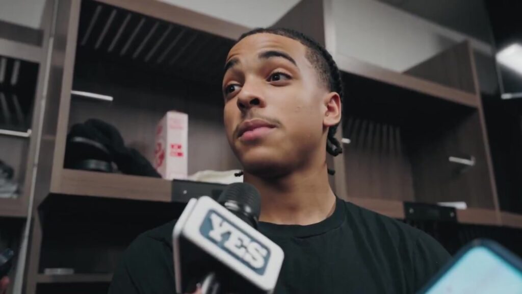 Mikel Brown Jr. in a black T-shirt speaking to reporters in the visitors' locker room in Boston