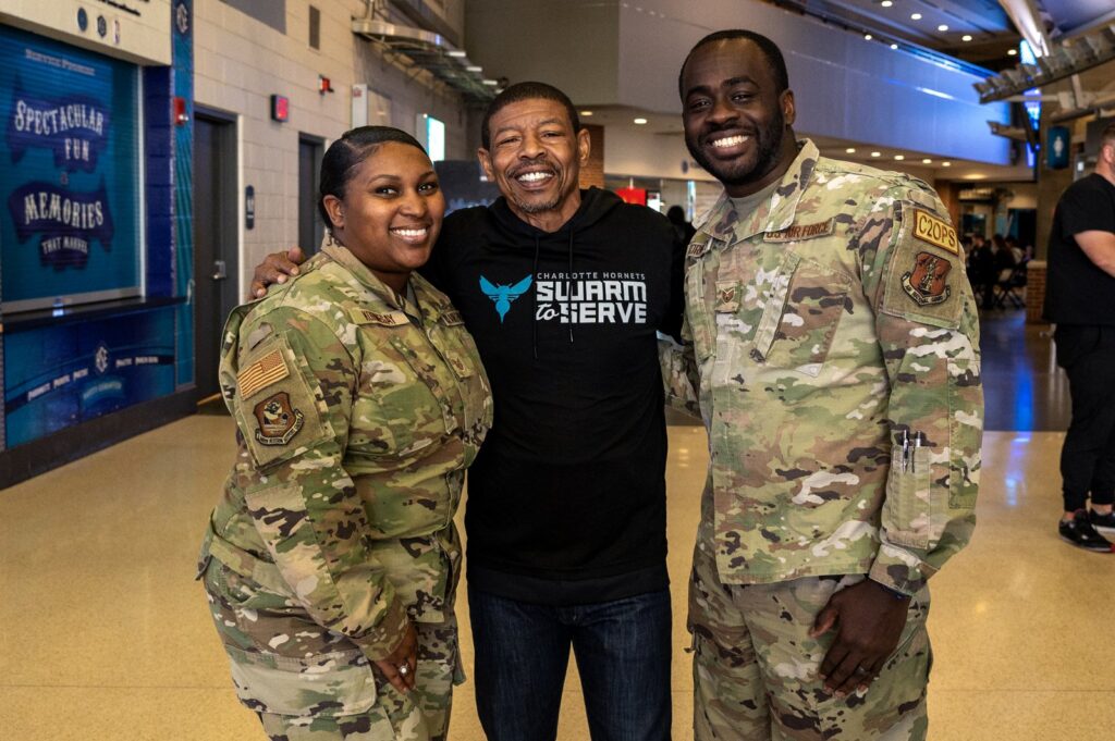 Muggsy Bogues smiles with two U.S. service members at Spectrum Center in Charlotte, January 2025