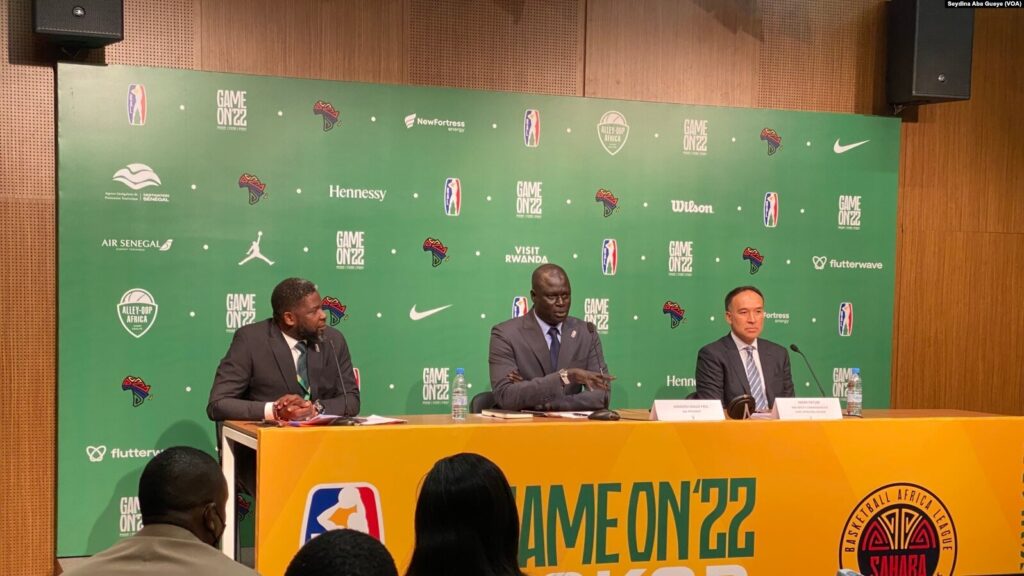 NBA deputy commissioner Mark Tatum, right, seated at a press conference table with two other officials in front of a green sponsor backdrop