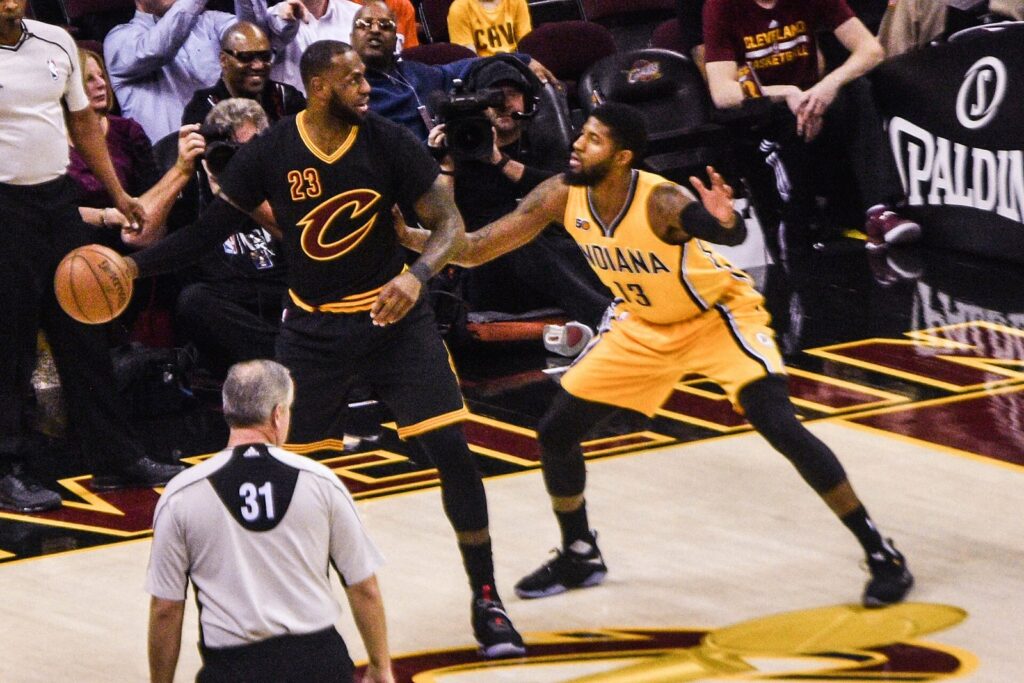 Paul George in an Indiana Pacers uniform guarding LeBron James as a referee watches