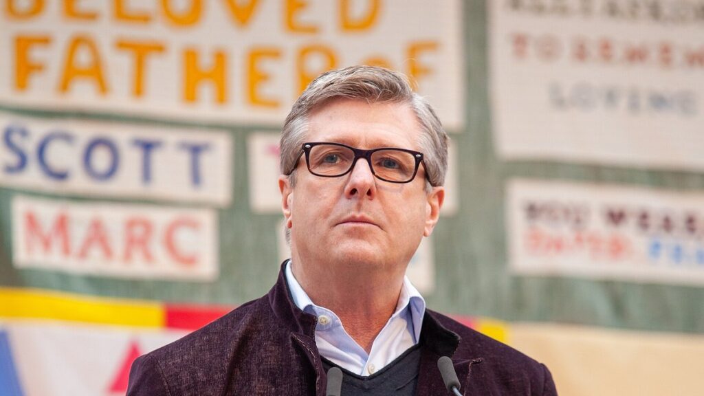 Rick Welts speaks at a podium in front of memorial quilt panels in San Francisco